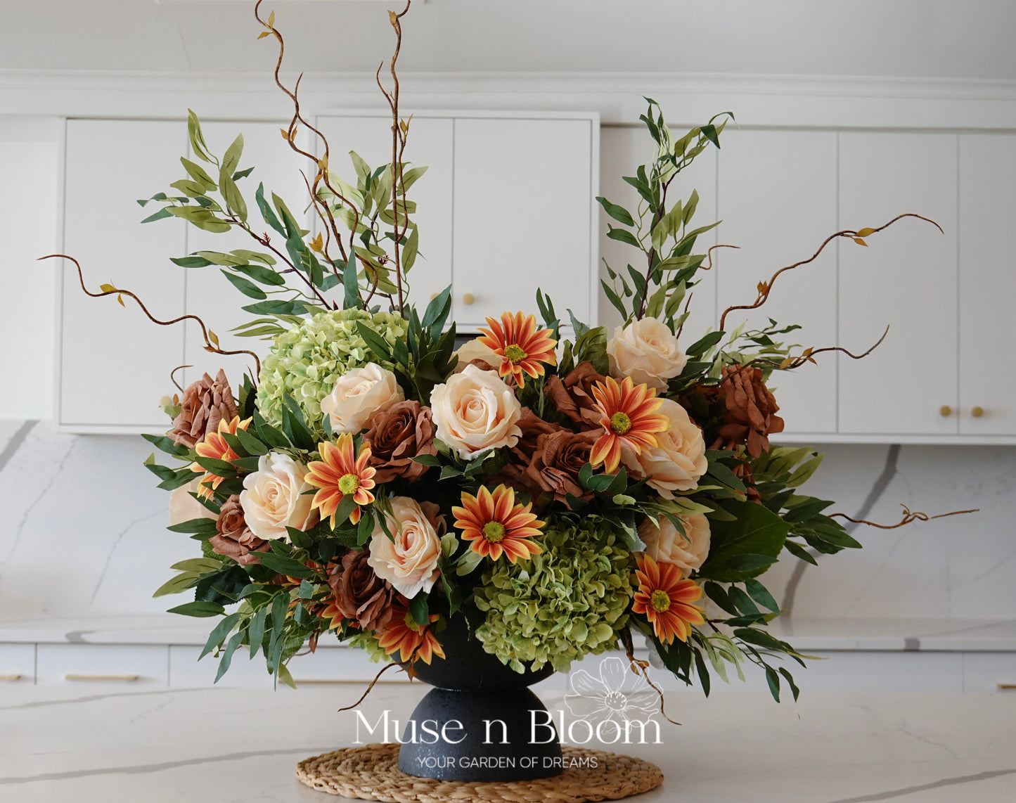 Floral arrangement with greenery and colorful flowers on a kitchen counter, featuring the brand 'Muse n Bloom'.